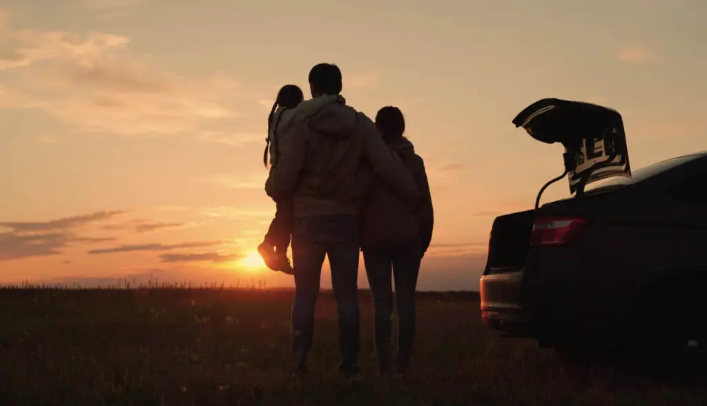 Father, wife, and daughter silhouetted against sunset, standing by car trunk symbolizing faith, family, and fatherhood.